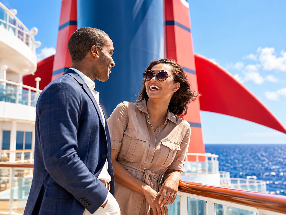 A couple laughs together on a cruise ship deck beside the ship's red funnel, with the open sea behind them