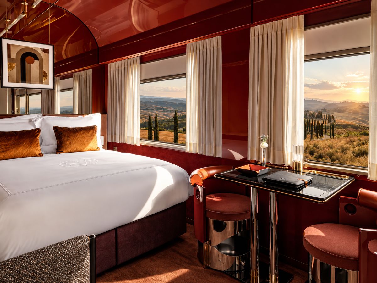 A luxurious private train suite with a plush bed and dining table, overlooking the rolling hills and cypress trees of the Tuscan countryside at sunset